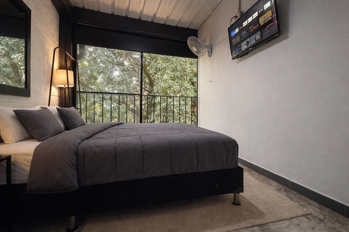 Quarto com uma cama, televisão na parede e janela com vista para árvores.