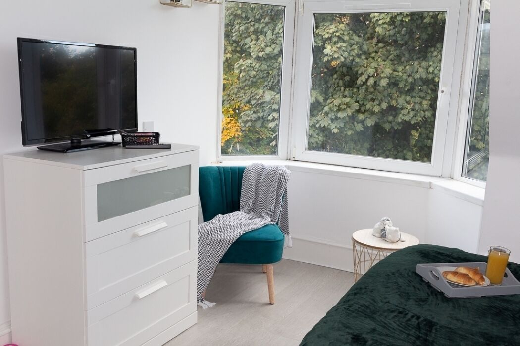 A room with a bed, a TV on a dresser, a chair with a blanket, a small table with a cup, and a window with a view of trees.