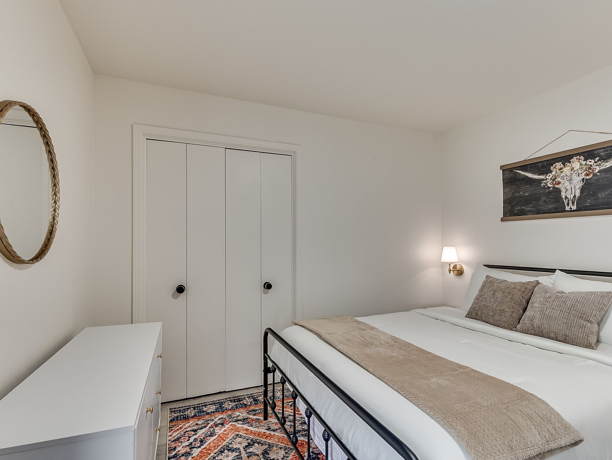 Ein Schlafzimmer mit einem Bett, einem Nachttisch, einer Tür, einer Wandleuchte und einem Bild.