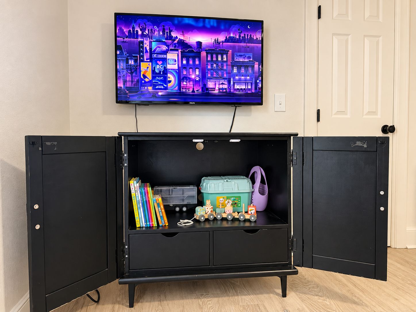A room with a television mounted on the wall displaying a cityscape. In the center, there is a black cabinet with open doors revealing books, a toy train, and a green container.