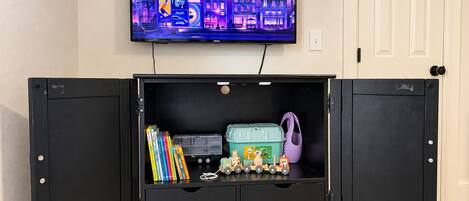 A room with a television mounted on the wall displaying a cityscape. In the center, there is a black cabinet with open doors revealing books, a toy train, and a green container.