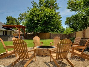 Ein Garten mit einem Feuerkorb, umgeben von Holz-Adirondack-Stühlen, einer überdachten Terrasse und einer Ziegelwand.