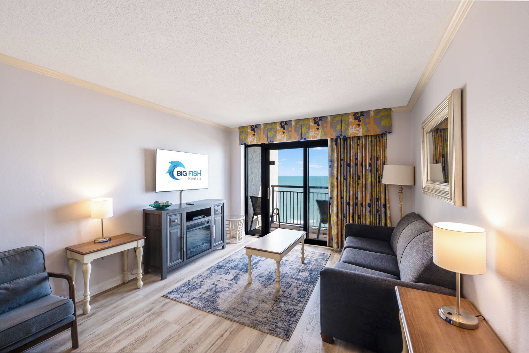 Ein Hotelzimmer mit Flachbildfernseher, einer Couch, einem Couchtisch, einem Fernsehschrank, einem Balkon mit Vorhängen und Blick aufs Meer.