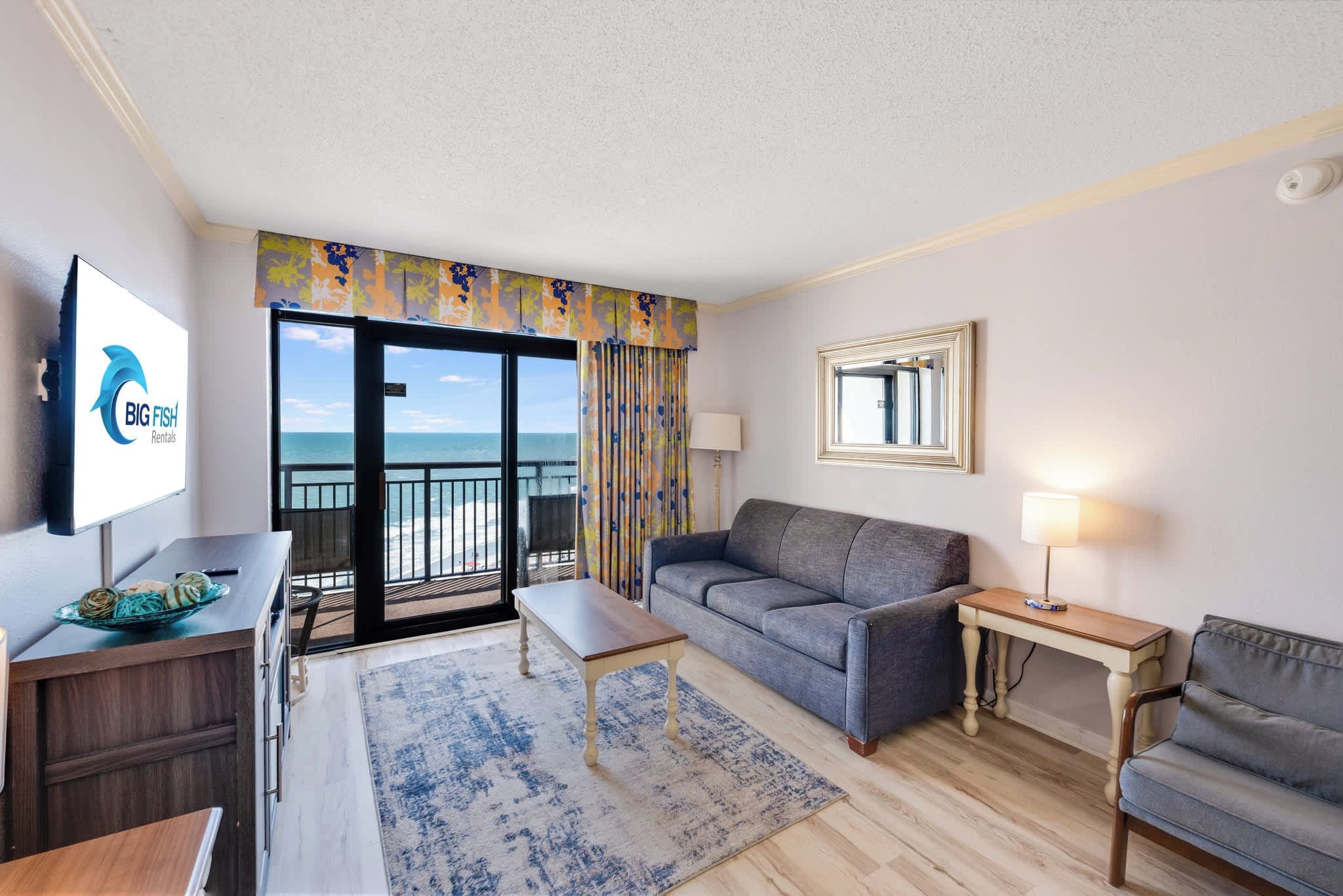A hotel room with a balcony, a sofa, a table, a lamp, and a television displaying the Big Fish Rentals logo.