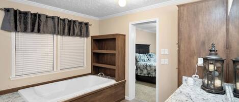 A bathroom with a bathtub, a sink, and a vanity. There is a window with blinds, a door leading to a bedroom, and a ceiling light.