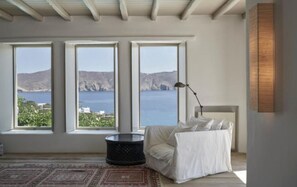 A room with a large window overlooking a coastal landscape, a white sofa, a floor lamp, and a patterned rug. - Villa Lioren, Divine Sea Views, Private Pool, Furnished Terrace, Mykonos (Mykonos)