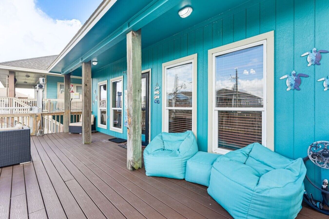 A covered deck with a blue exterior, wooden flooring, and a light blue cushioned seating area.