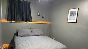 A bedroom with a bed, a headboard, a window with curtains, a wall-mounted picture, and a small bedside table.