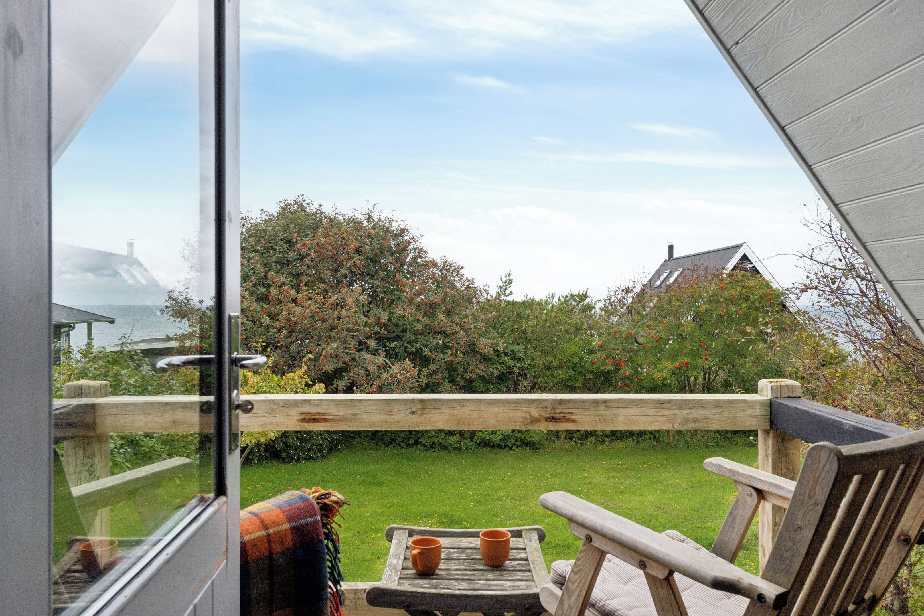 Een houten terras met een tafel, stoelen, een glazen deur en uitzicht op een tuin en gebouwen.