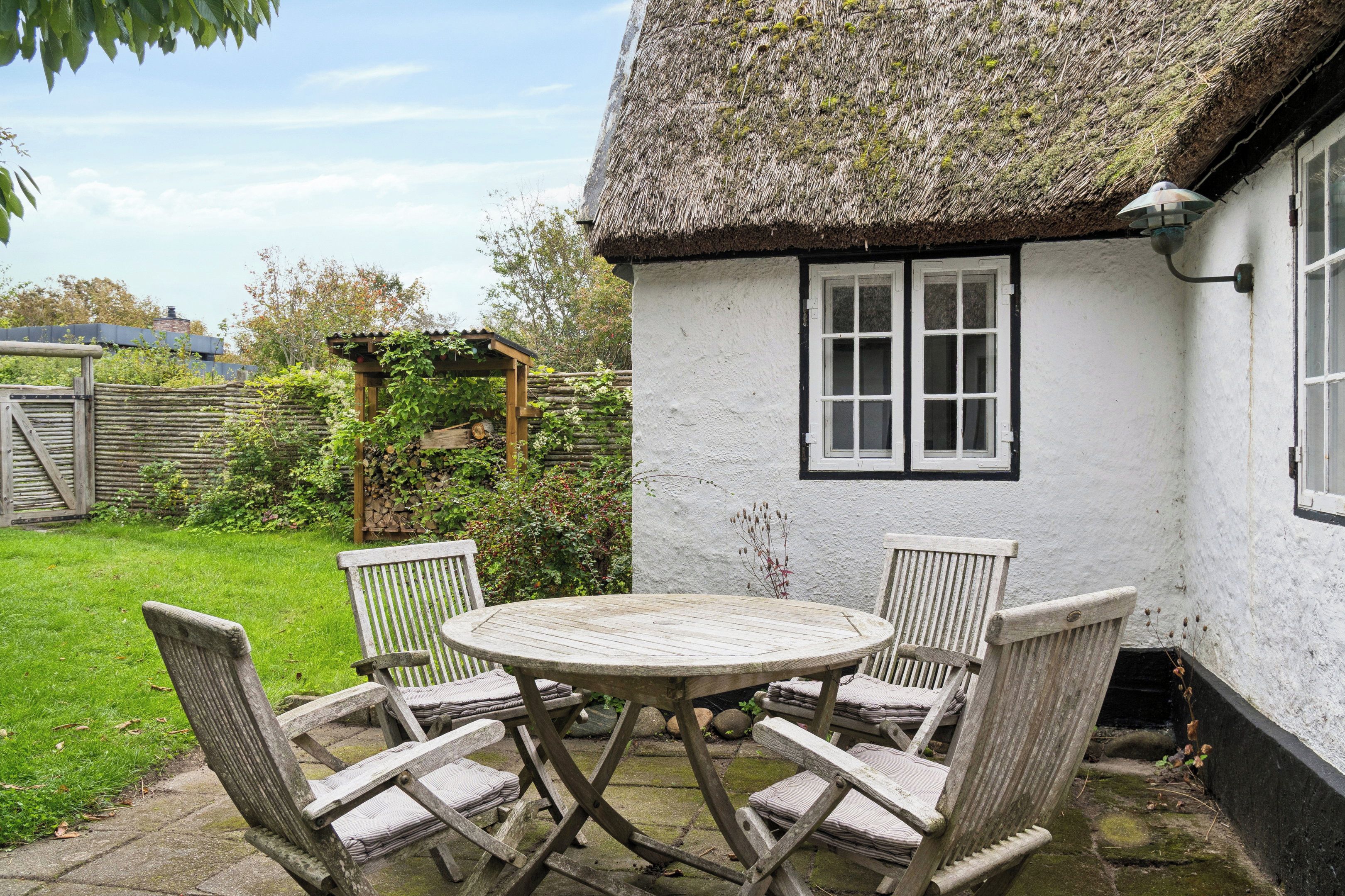 Une petite maison au toit de chaume, à l’extérieur blanc, avec une table ronde en bois et quatre chaises, ainsi qu’un petit jardin.