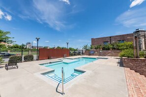 Una piscina rectangular con una línea de agua azul cristalina, rodeada por una terraza pavimentada. Hay sillones de descanso y una silla de ruedas cerca de la piscina. Se observa un edificio de ladrillo con fachada de piedra al fondo.