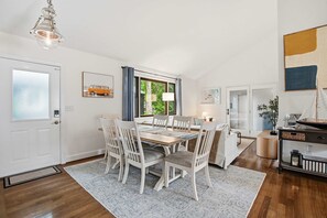 Ein Essbereich mit einem Holztisch und weißen Stühlen, einem Fenster mit Vorhängen sowie einer hängenden Leuchte.