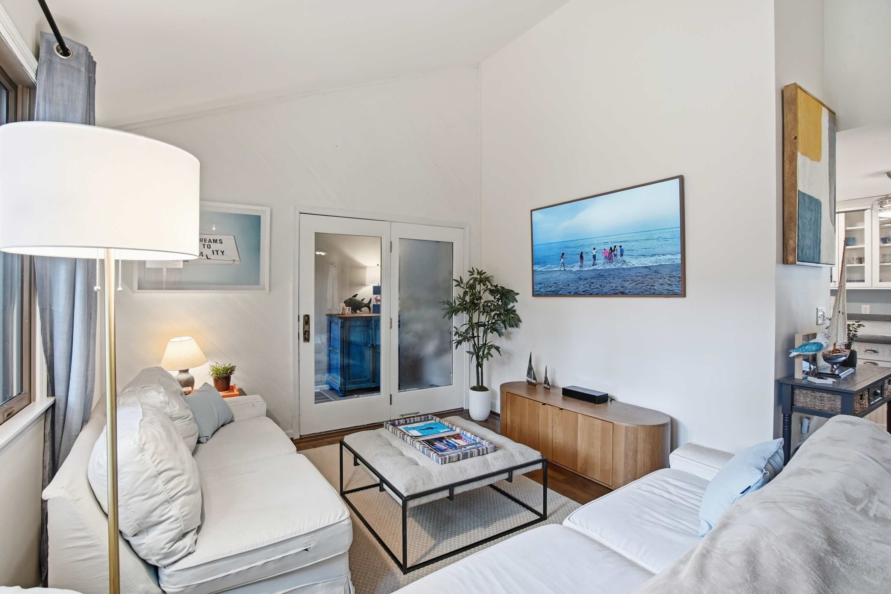 Une chambre d’hôtel moderne, dotée d’un canapé, d’une table basse, d’une bibliothèque, d’une plante en pot et d’une grande fenêtre donnant sur la plage.