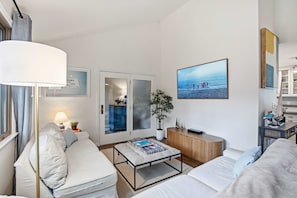 Ein modernes Hotelzimmer mit einer Couch, einem Couchtisch, einem Bücherregal, einem Topf mit Pflanze und einem großen Fenster mit Blick auf den Strand.