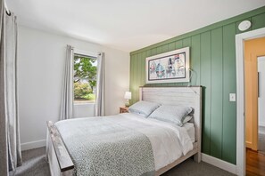 Ein Schlafzimmer mit einem Bett, einem fensterbelaubten Fenster, einer Nachttischlampe und einem gerahmten Bild an der Wand.