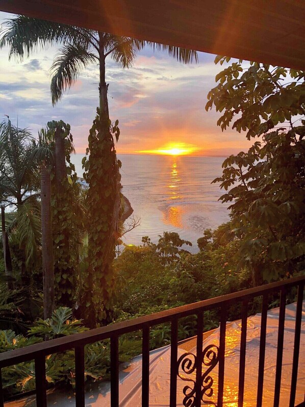A balcony with a view of a sunset over the ocean, surrounded by tropical vegetation. - The King's Panoramic Ocean View (Unknown)