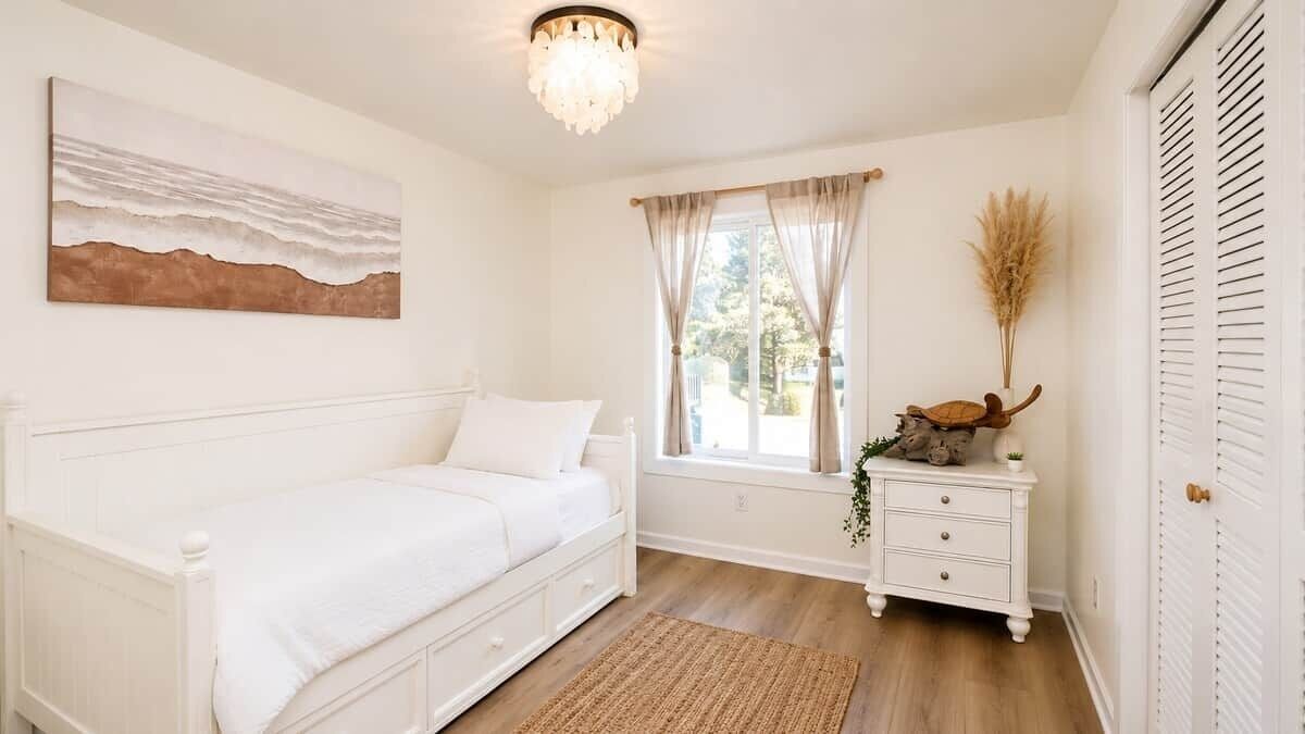 Een slaapkamer met een wit bed, een nachtkastje met een plant, een raam met gordijnen en een aan de muur hangend schilderij.