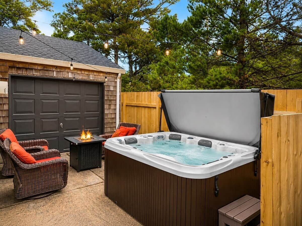 A hot tub with a wooden deck, a wicker chair with a red cushion, and a table with a fire feature.