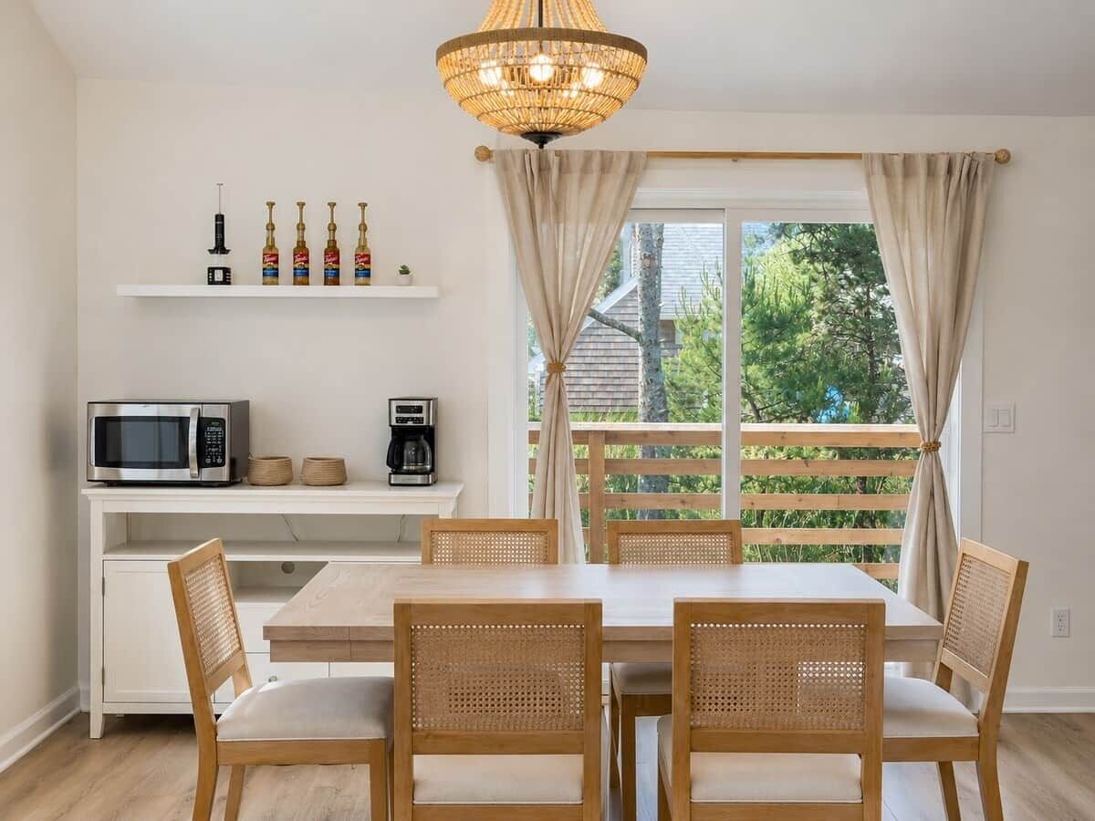 A dining area with a white table, four wicker chairs, a white cabinet with kitchen appliances, and a large window with curtains.