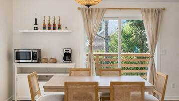 A dining area with a white table, four wicker chairs, a white cabinet with kitchen appliances, and a large window with curtains.