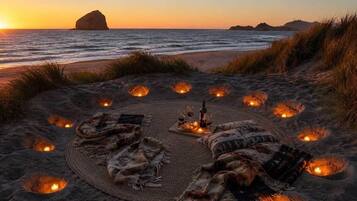 A beach setting with a circular arrangement of candles and blankets, suggesting a romantic or intimate outdoor dining experience.