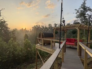 A wooden deck with a red sofa, a barrel, and hanging lights. - Scarlet Ember - Hot Tub, Barrel Sauna, Sunset Views, & King Beds! (Campton)