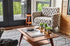 A modern living room with a patterned armchair, a coffee table with a remote control, and potted plants. - Scarlet Ember - Hot Tub, Barrel Sauna, Sunset Views, & King Beds! (Campton)