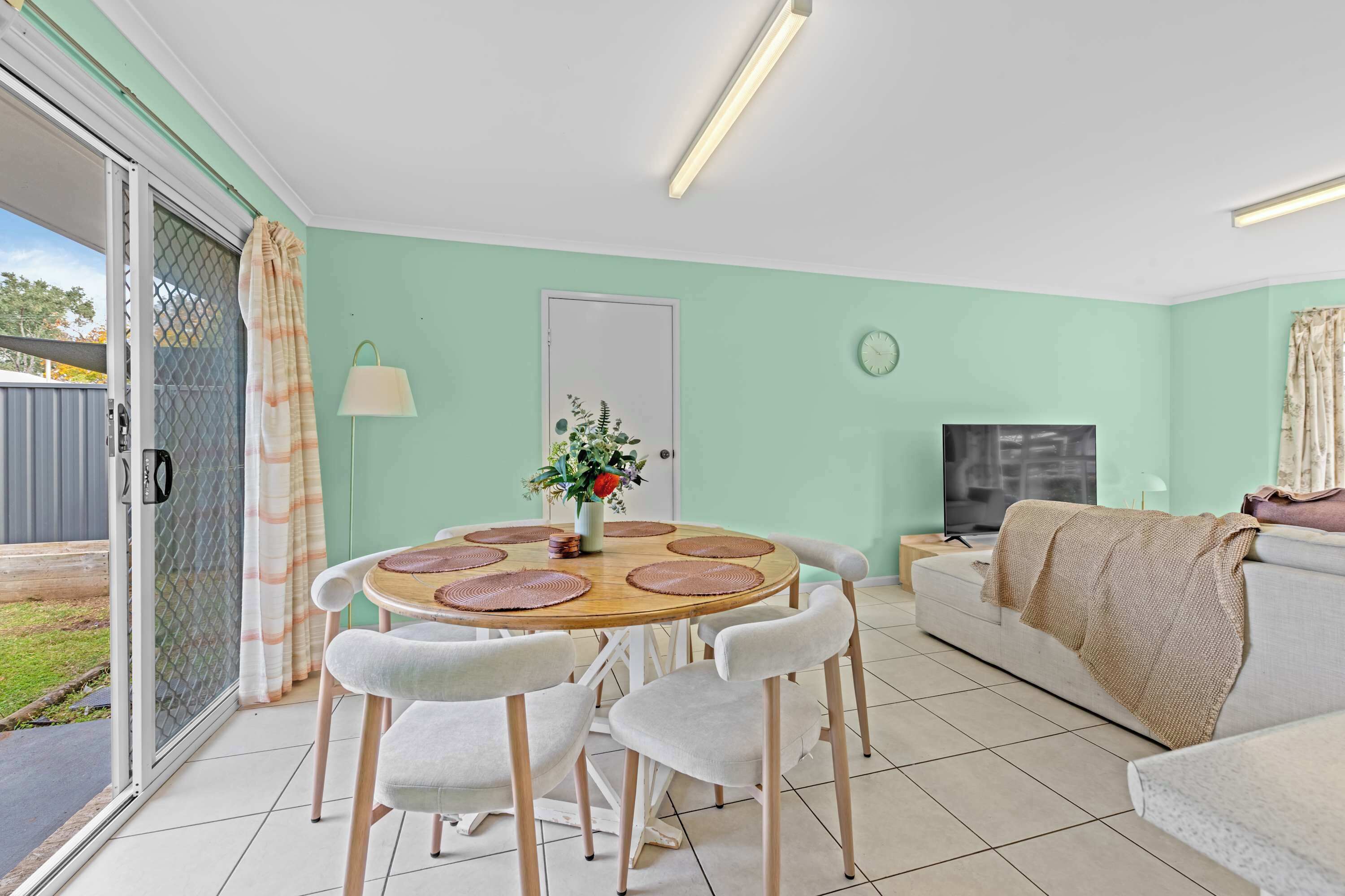 Un coin repas avec une table ronde, des chaises blanches, un vase de fleurs et un téléviseur en arrière-plan.