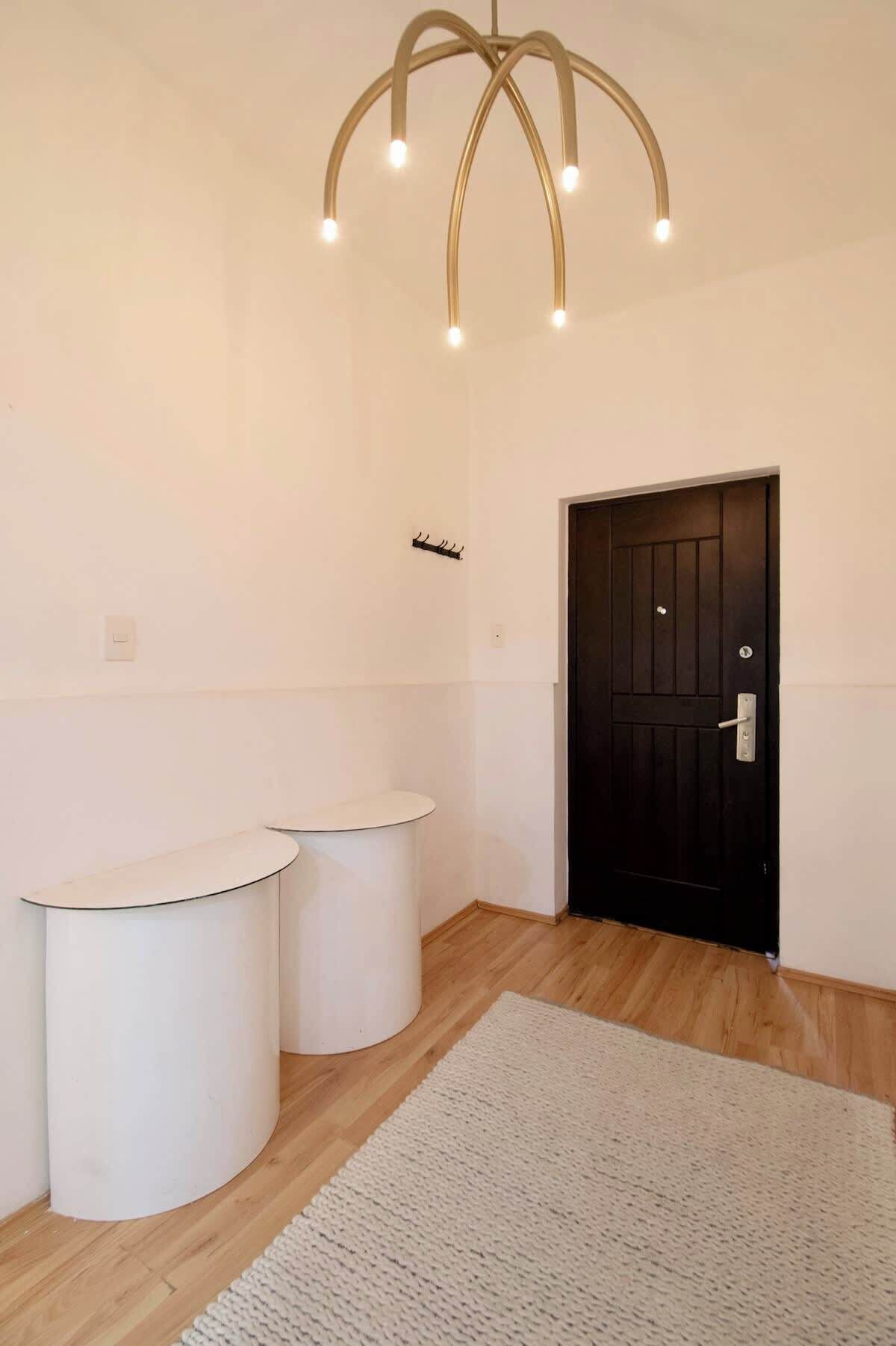 A room with a black door, two white cylindrical stools, a white rug, and a modern light fixture.