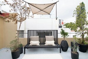 En sort metalbænk med hynder, omgivet af potteplanter, under et solsejl på en tagterrasse.