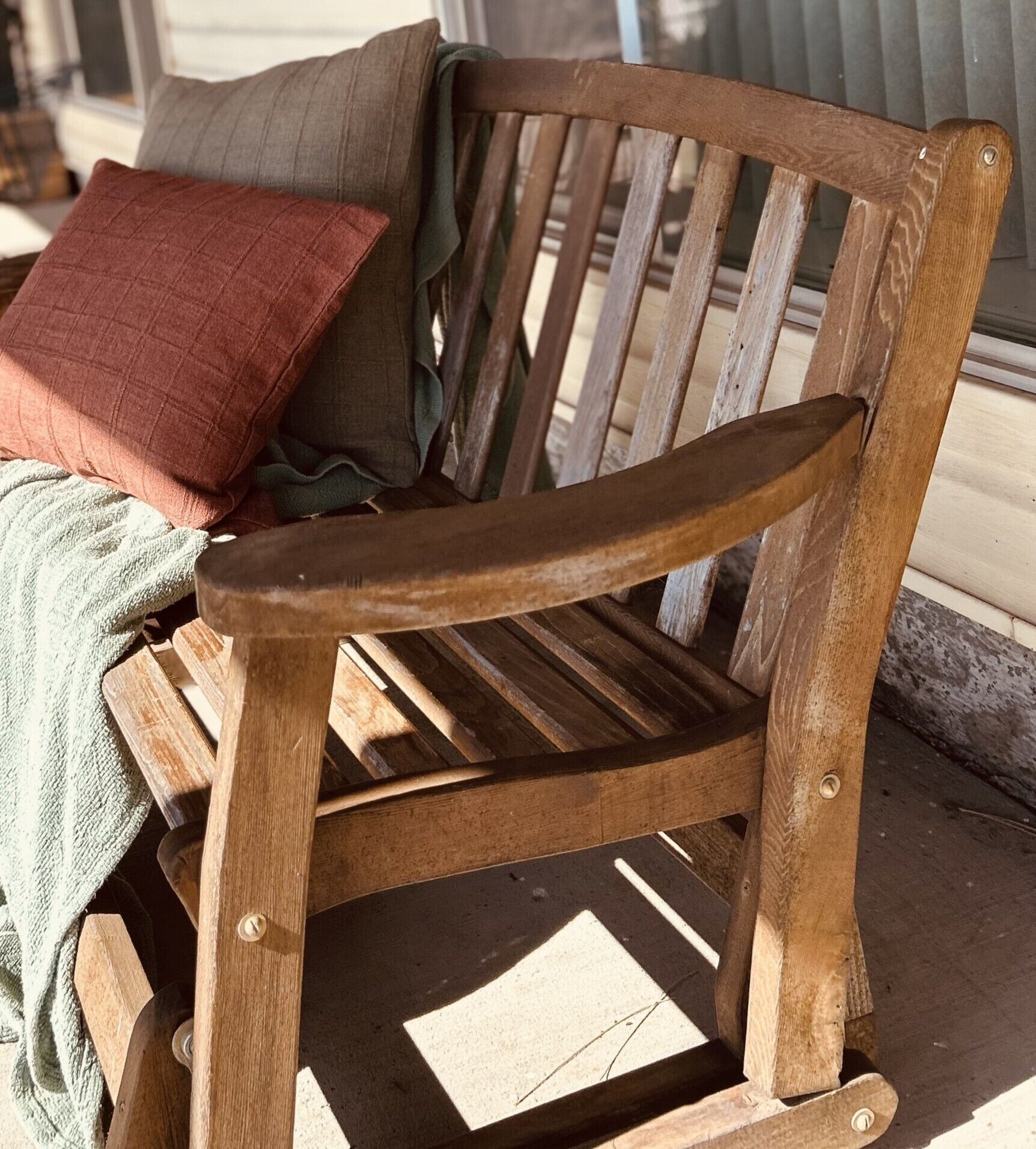 A wooden bench with a slatted backrest and armrests, positioned on a porch with a cushion and a blanket.