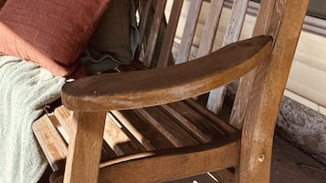 A wooden bench with a slatted backrest and armrests, positioned on a porch with a cushion and a blanket.