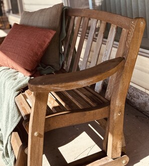 A wooden bench with a slatted backrest and armrests, positioned on a porch with a cushion and a blanket.