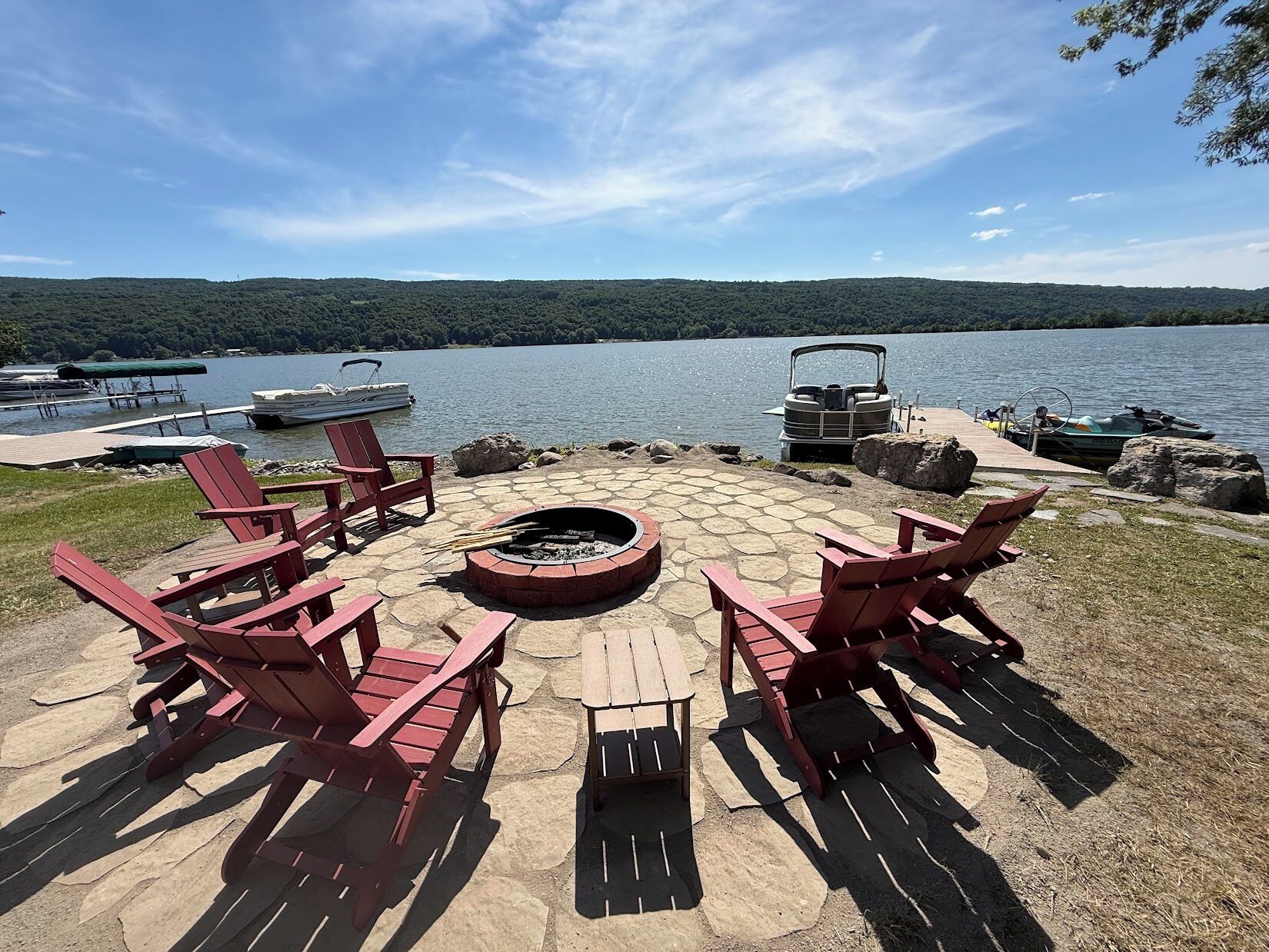 A lakeside deck with Adirondack chairs, a fire pit, and boats docked in the background.
