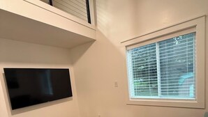 A living room with a flat-screen TV mounted on the wall, a window with blinds, a brown leather ottoman, and a wooden floor. - Newly Renovated Family-Friendly 3-Bedroom Otisco Lake Home (Marietta)