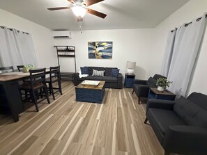 Una sala de estar moderna con piso de madera, ventilador de techo, comedor con mesa y sillas, y un juego de sofás.