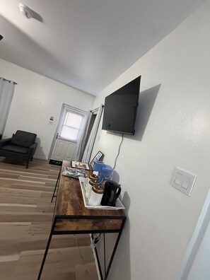 Ein Wohnzimmer mit einem an der Wand befestigten Flachbildfernseher, einem Couchtisch aus Holz mit diversen Gegenständen, einem dunkelgrauen Sofa und einer Tür mit Fenster.