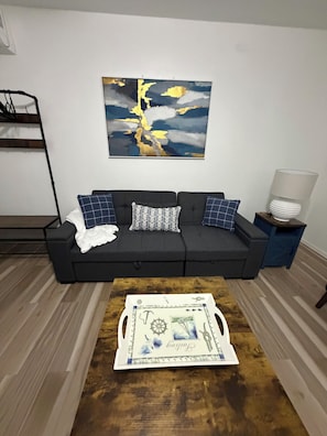 Ein Wohnzimmer mit schwarzem Sofa, gemusterten Kissen, einem Couchtisch mit dekorativer Ablage, einem Wandbild und einem Beistelltisch mit Lampe.
