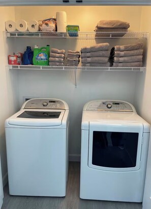 A laundry room with two front-loading washing machines and shelves above containing towels, toilet paper, and cleaning supplies.