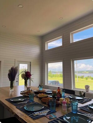 A dining area with a long table set for a meal, large windows with a view of a lake, and a door leading outside.