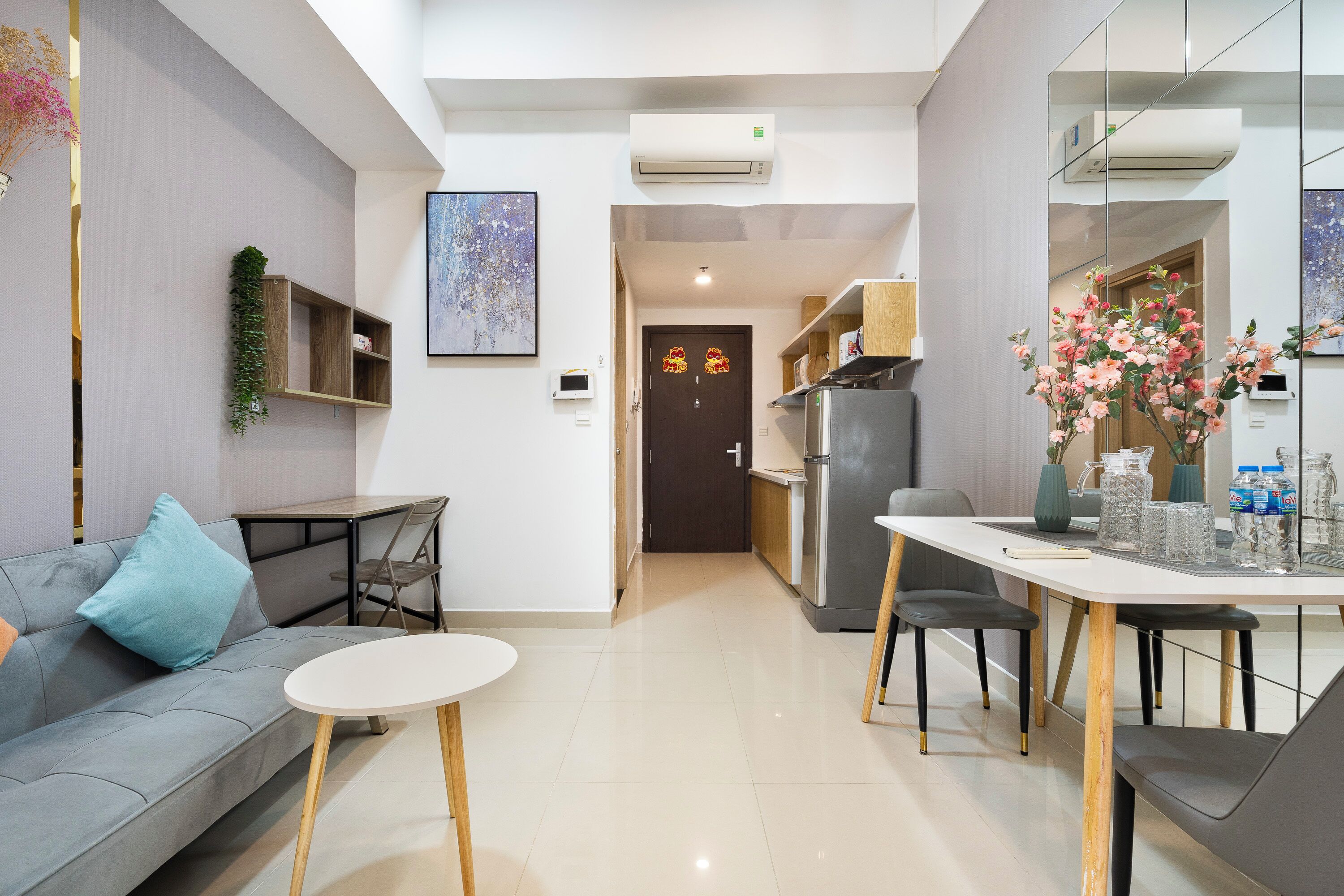 Uma sala de estar moderna com um sofá cinzento, uma mesa redonda pequena e uma cozinha com frigorífico e mesa de jantar.