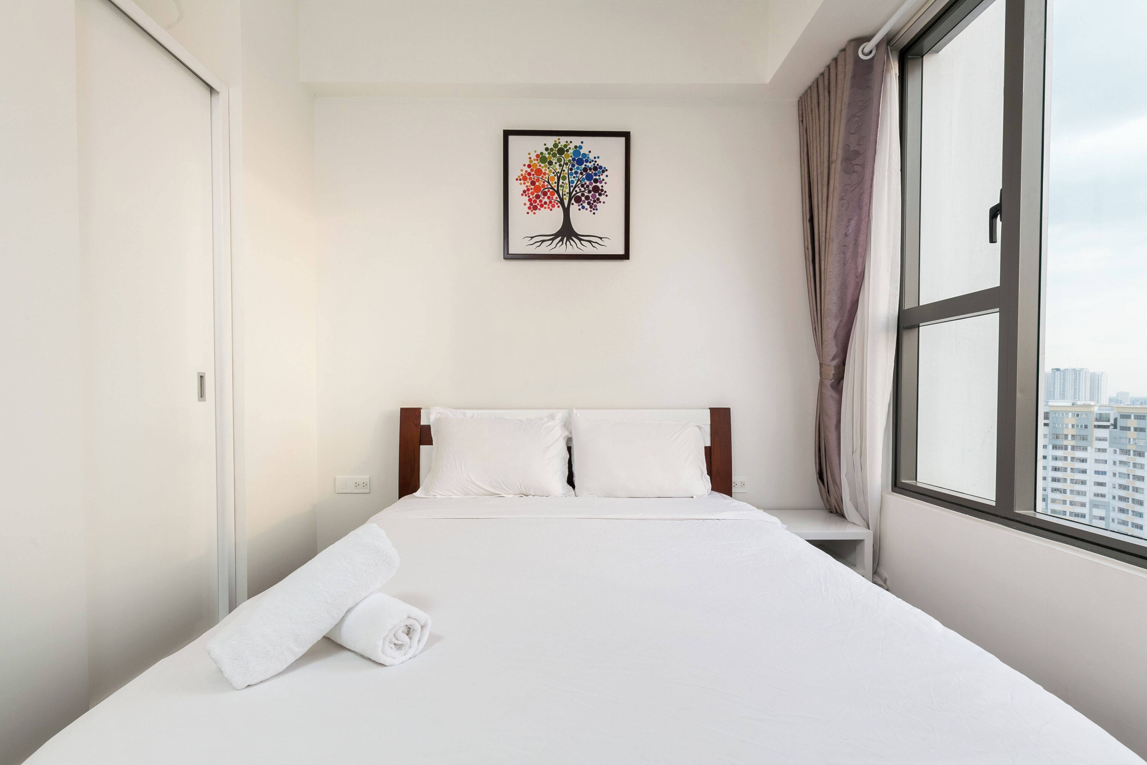 A hotel room with a bed, a window with curtains, and a wall-mounted picture of a tree.