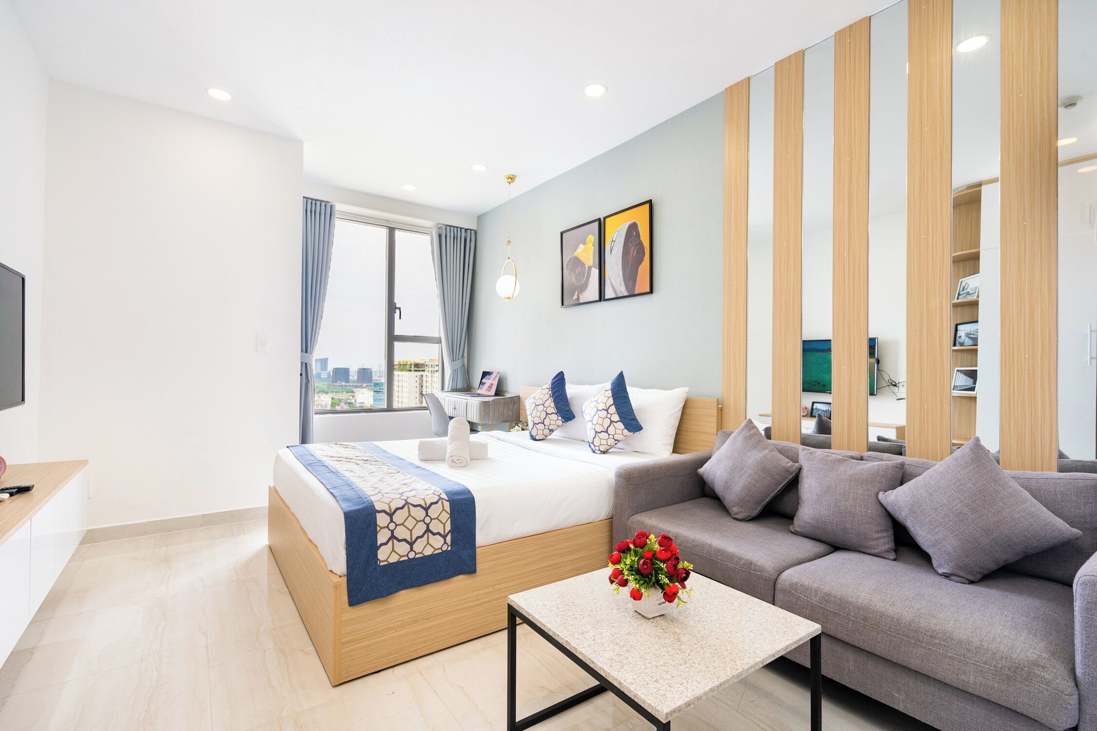 Una habitación de hotel moderna con cama, sofá, una mesita con un jarrón de flores y vistas a la ciudad por la ventana.