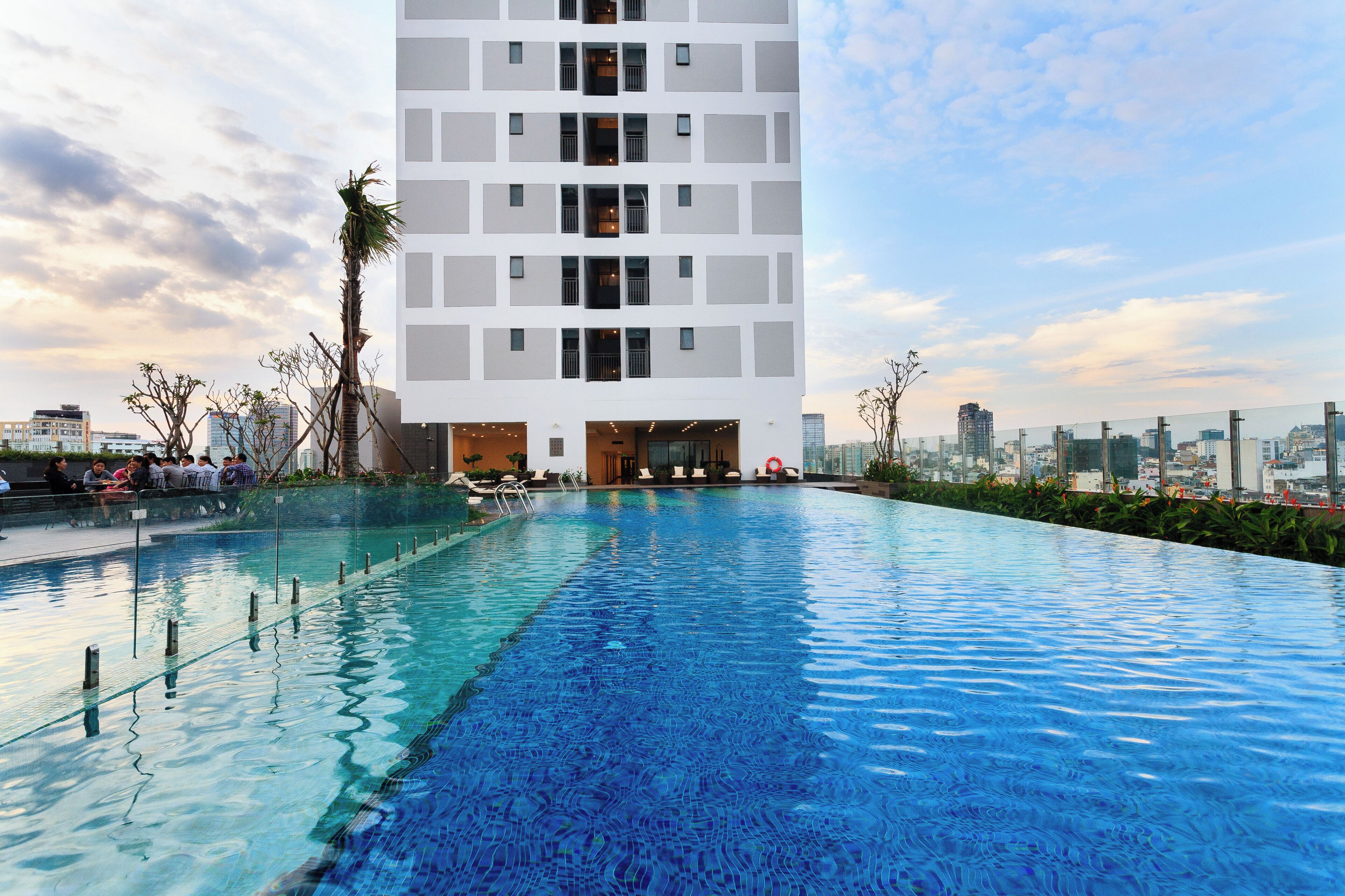 Um edifício moderno com piscina no terraço, rodeado por vegetação e uma paisagem urbana ao fundo.