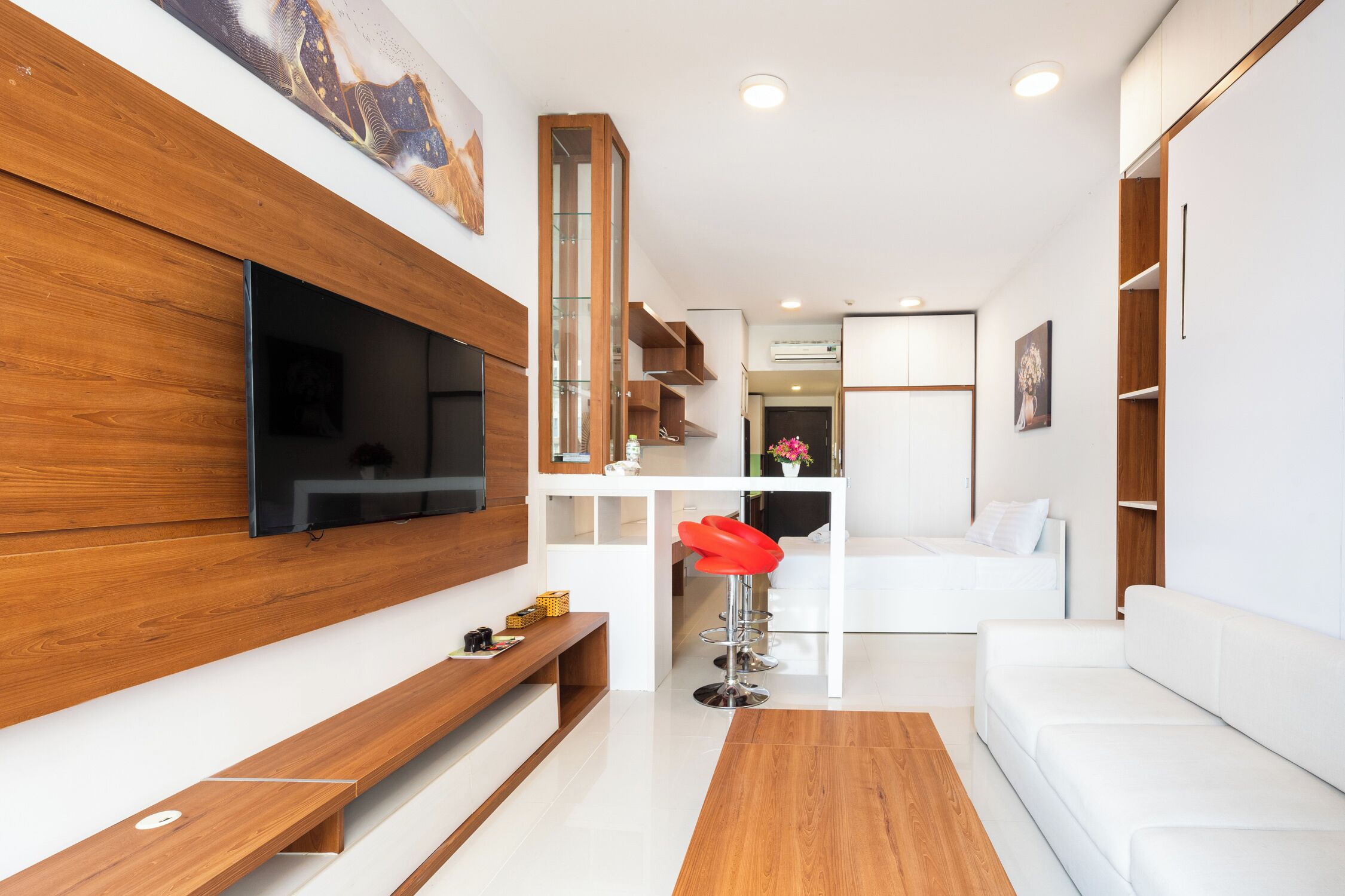 A modern living room with a flat-screen TV mounted on a wooden wall, a white sofa, a red bar stool, and a wooden coffee table.