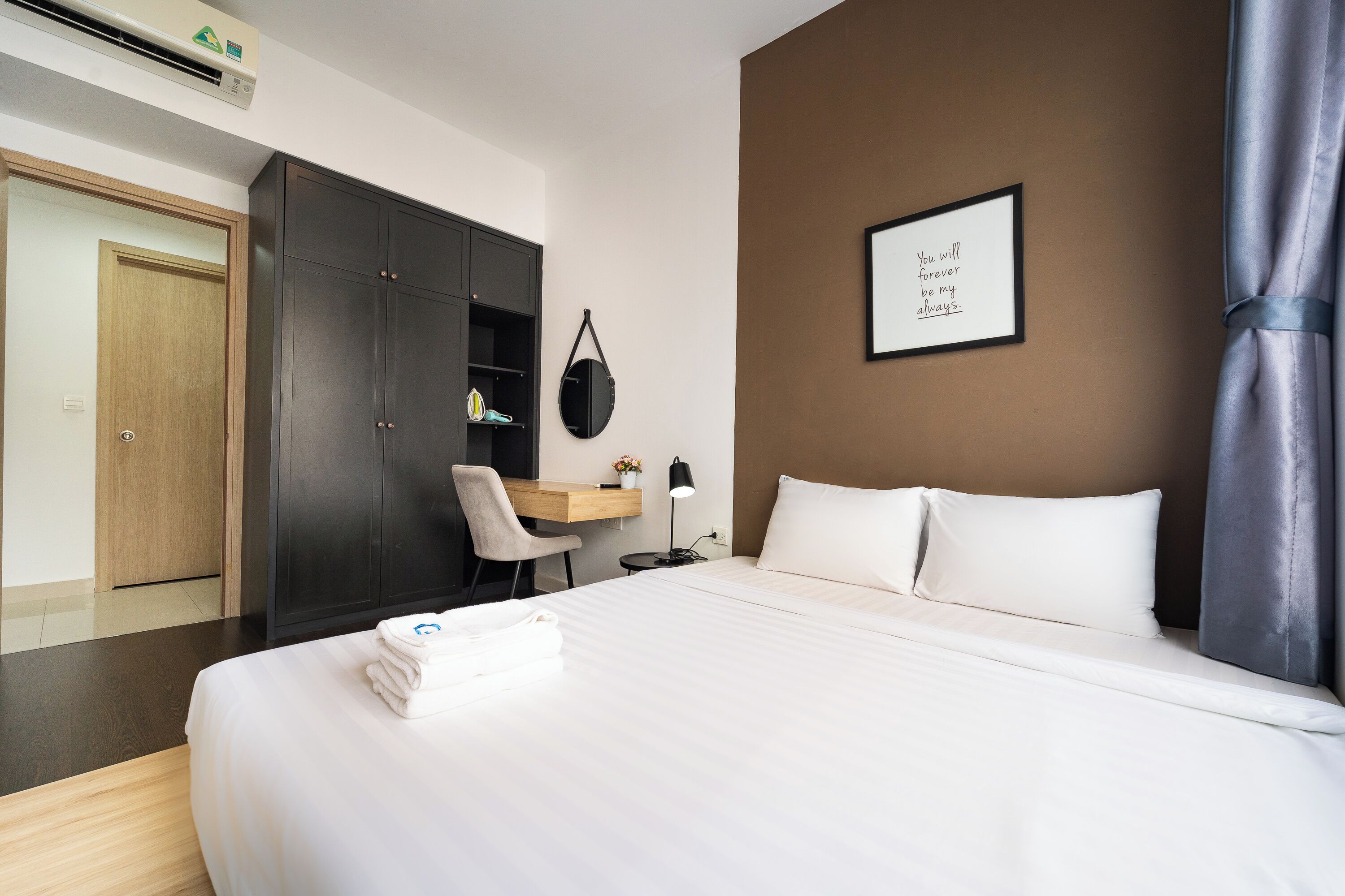 A hotel room with a bed, a desk, a chair, a wardrobe, and a framed quote on the wall.