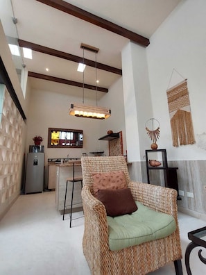 A cozy living area with a wicker chair, a kitchen in the background, and a hanging light fixture. - Beautiful apartment with a pool and VIP area on Paseo Montejo in Mérida, Yucatán (Mérida)