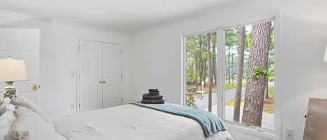 A bedroom with a bed, a lamp, a window with a view of trees, and a door.