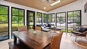 A spacious room with a large wooden table, comfortable seating, and large windows offering a view of the outdoors. - 3BR New Renovation, with pool, tennis, trolley! (Bethany Beach)