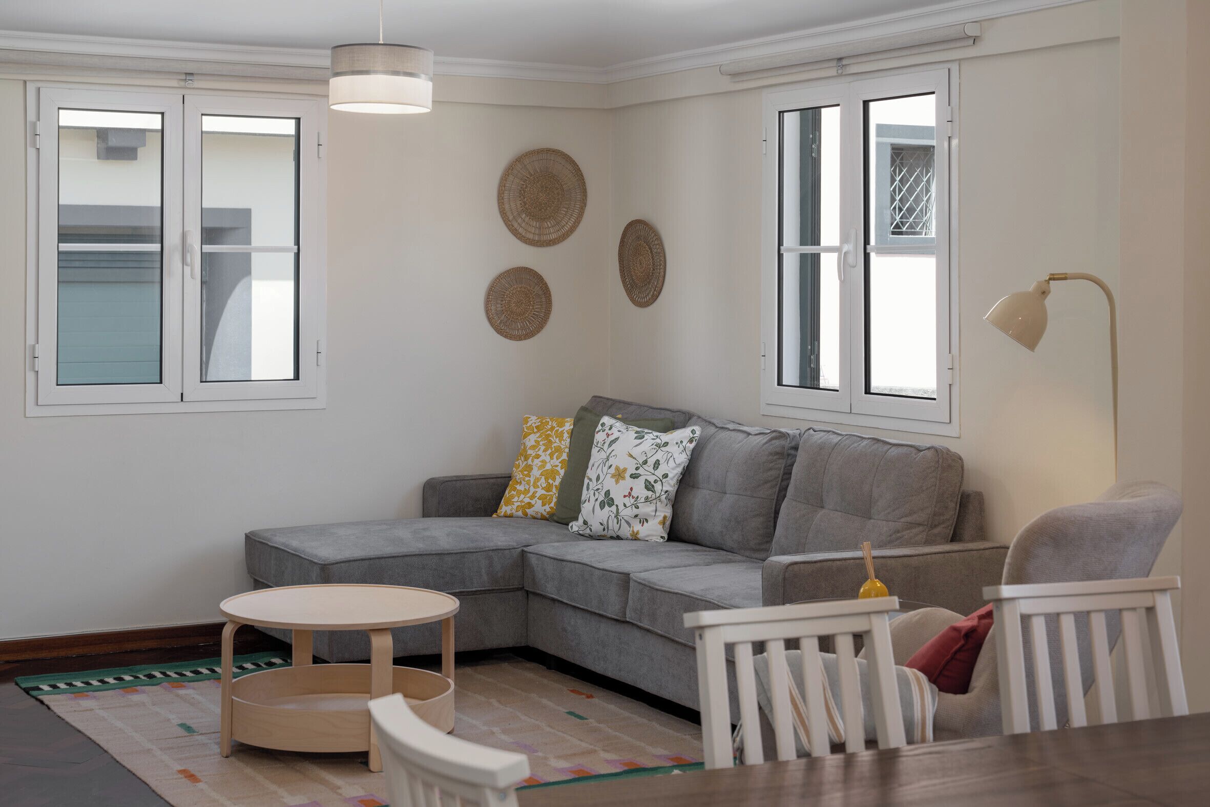 A modern living room with a grey sectional sofa, a round wooden table, and a white armchair. There are three windows with white frames, and a floor lamp is visible.